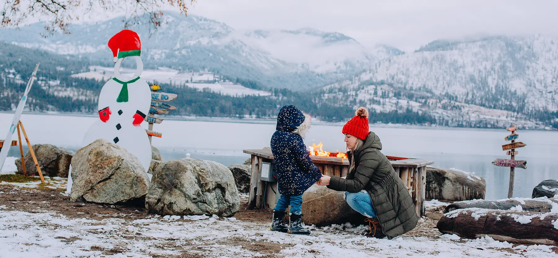 A parent and child at a lakeside firepit in winter, snow on the far hills